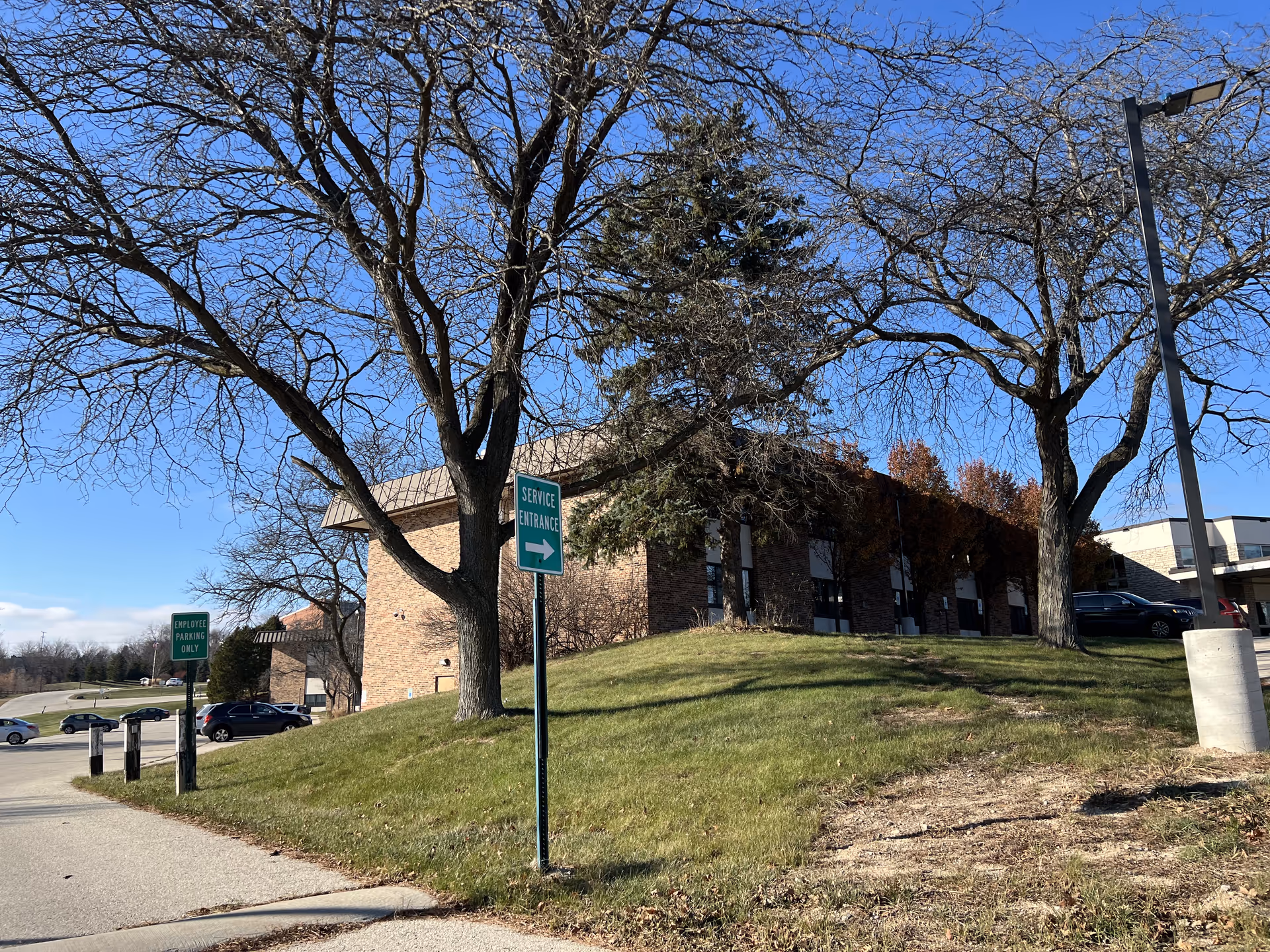 Exterior view of a brick building with leafless trees and a grassy hill in front. There are signs indicating 'Service Entrance' and 'Employee Parking Only' near a parking lot with several cars. The sky is clear and blue.
