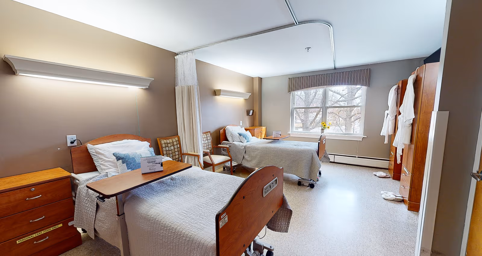 A bright and clean shared bedroom in a healthcare facility with two hospital-style beds, each with white bedding and pillows. There are wooden nightstands beside each bed, two chairs between the beds, a large window with a view of trees, and wooden wardrobes with white robes hanging on the doors. The room has beige walls and a light-colored floor.