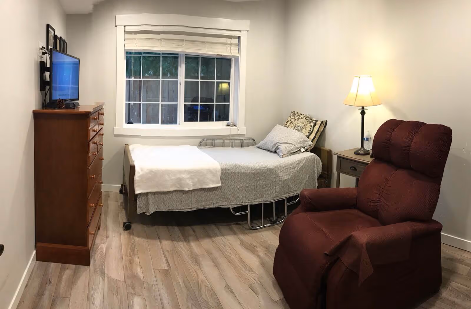 Furnished single-occupancy bedroom with a bed, red recliner, wooden dresser and TV, and a bedside lamp.