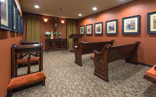 A small chapel room with wooden pews and kneelers upholstered in red fabric. The walls are painted a warm reddish-brown and decorated with framed religious artwork. At the front of the room, there is a wooden altar with floral arrangements and a crucifix mounted on the wall above it. The room is softly lit with recessed ceiling lights and wall sconces.