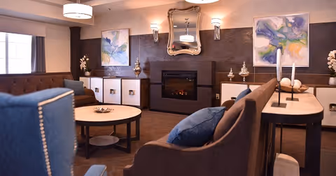A cozy living room area with brown and blue upholstered chairs and sofas arranged around a round coffee table. The room features a modern electric fireplace with a decorative mirror above it, flanked by two wall sconces. There are two abstract paintings on the walls and decorative vases on white cabinets beneath the paintings. The room is softly lit with ceiling lights and natural light from a window with curtains.