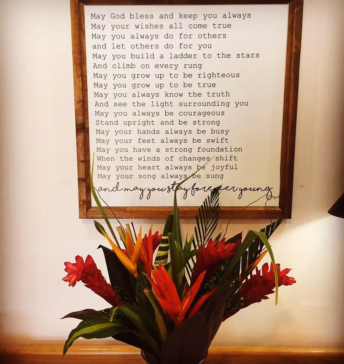 A framed inspirational poem with a wooden border hanging on a white wall above a colorful bouquet of tropical flowers with red and orange blooms and green leaves.