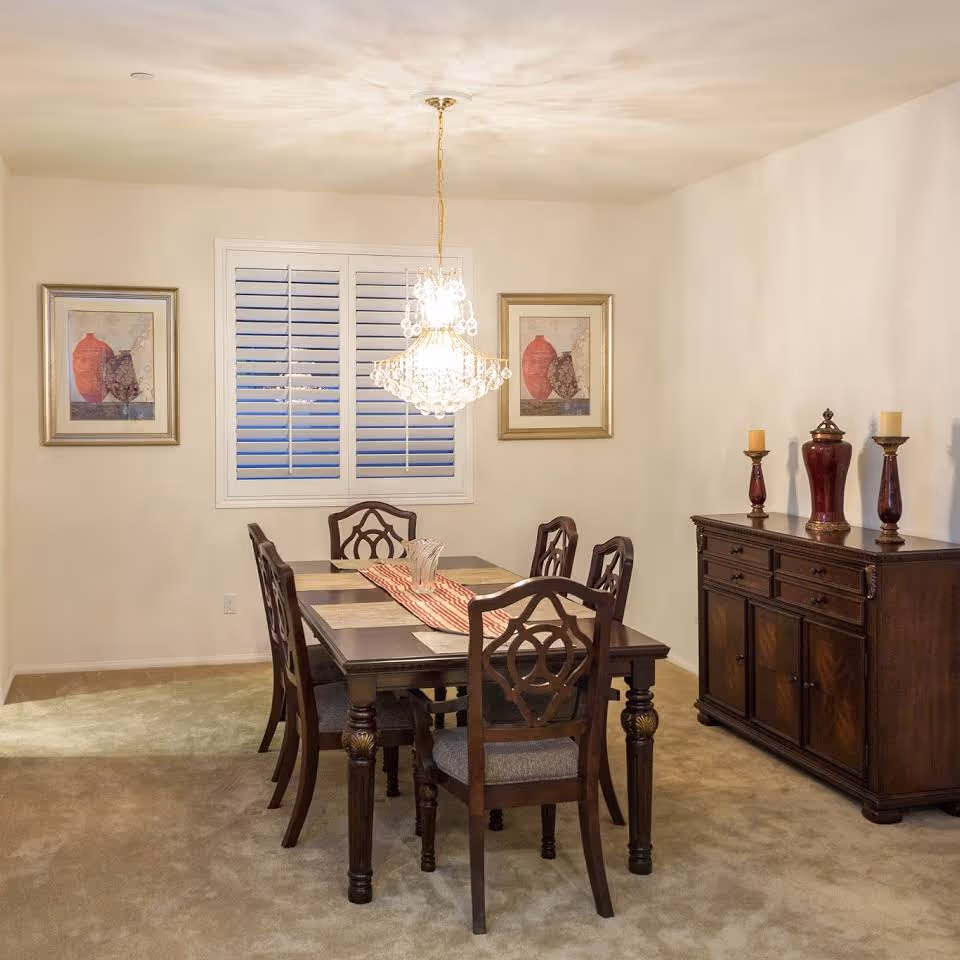 Traditional dining room with a wooden table and six chairs under a crystal chandelier, a sideboard with decorative vases, and framed artwork on the walls.