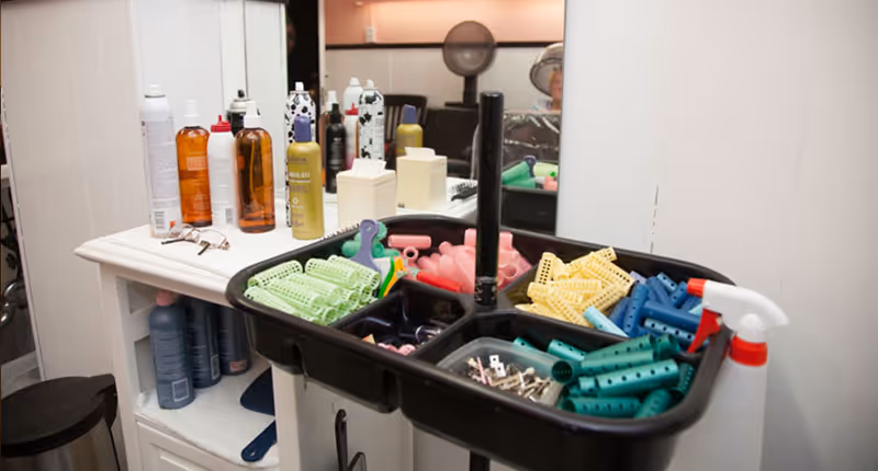 A salon station with a black tray holding various colored hair rollers and clips in front of a mirror. On the counter behind the tray, there are multiple bottles of hair care products and a pair of glasses. The background shows part of a salon chair and a fan.