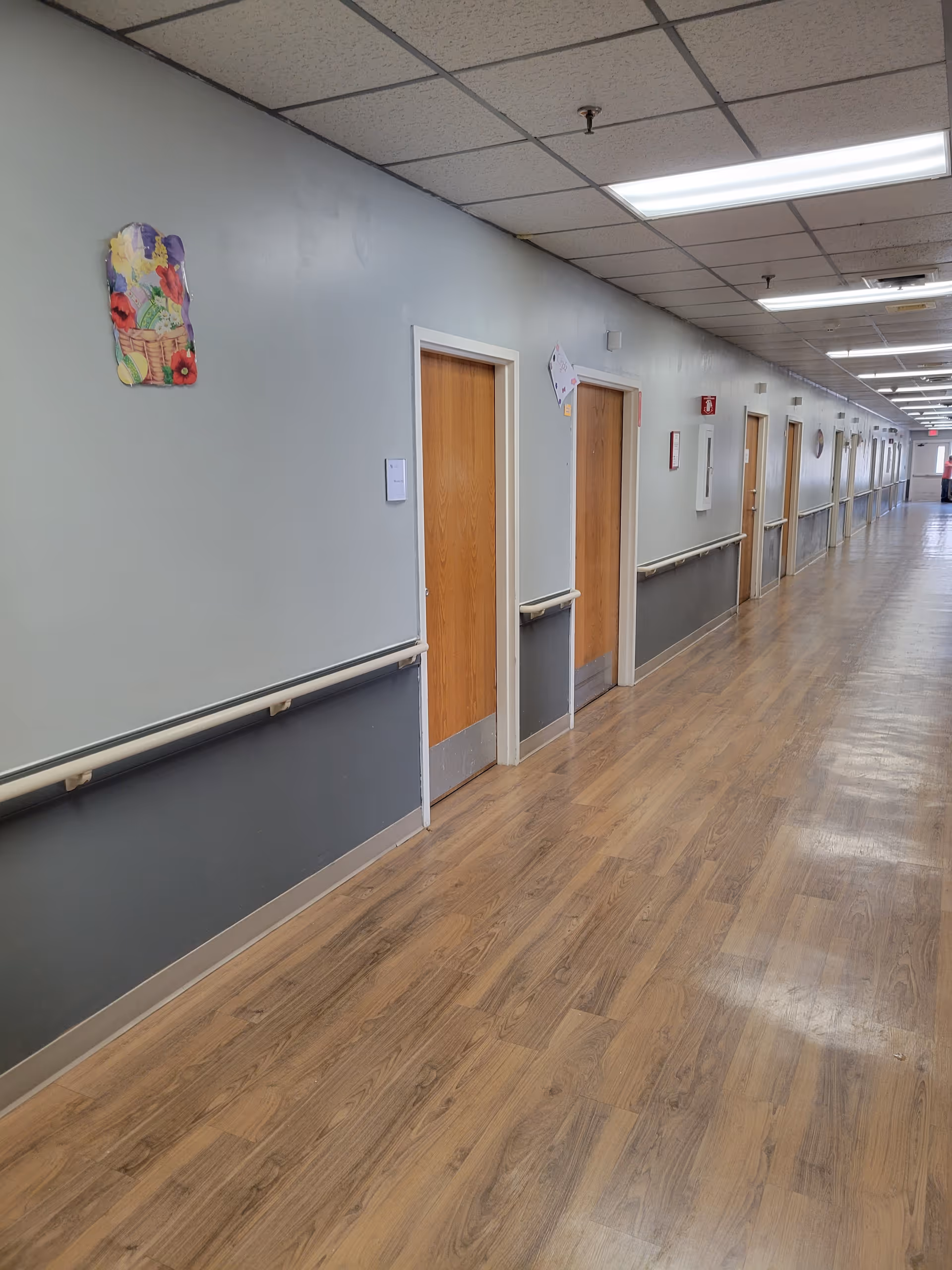 A long indoor hallway in an assisted living facility with wooden doors on the left side, handrails along the walls, and a wood-patterned floor. The ceiling has fluorescent lights and a drop ceiling design. Some decorations are visible on the walls.