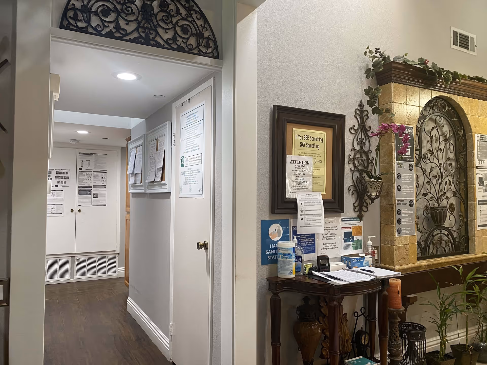 Interior hallway of Cypress Residence featuring a small wooden table with hand sanitizer, disinfecting wipes, and informational signs about health and safety. The wall behind the table has decorative wrought iron wall hangings and framed notices. The hallway has wooden flooring and white walls with additional posted notices further down the corridor.