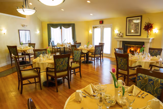 A warm and inviting dining room with several round tables covered in yellow tablecloths, each set with glassware, napkins, and silverware. The room features wooden chairs with patterned cushions, a lit fireplace with a framed picture above it, and large windows with curtains allowing natural light to enter.