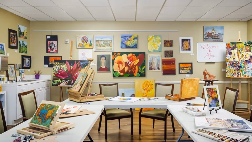 An art room with several tables arranged in a U-shape, each table holding painting supplies, canvases, and easels. The walls are decorated with various colorful paintings and artwork. There are chairs around the tables, and a whiteboard on the wall reads 'Welcome to Artna!'. The room has a wooden floor and a ceiling with fluorescent lighting.