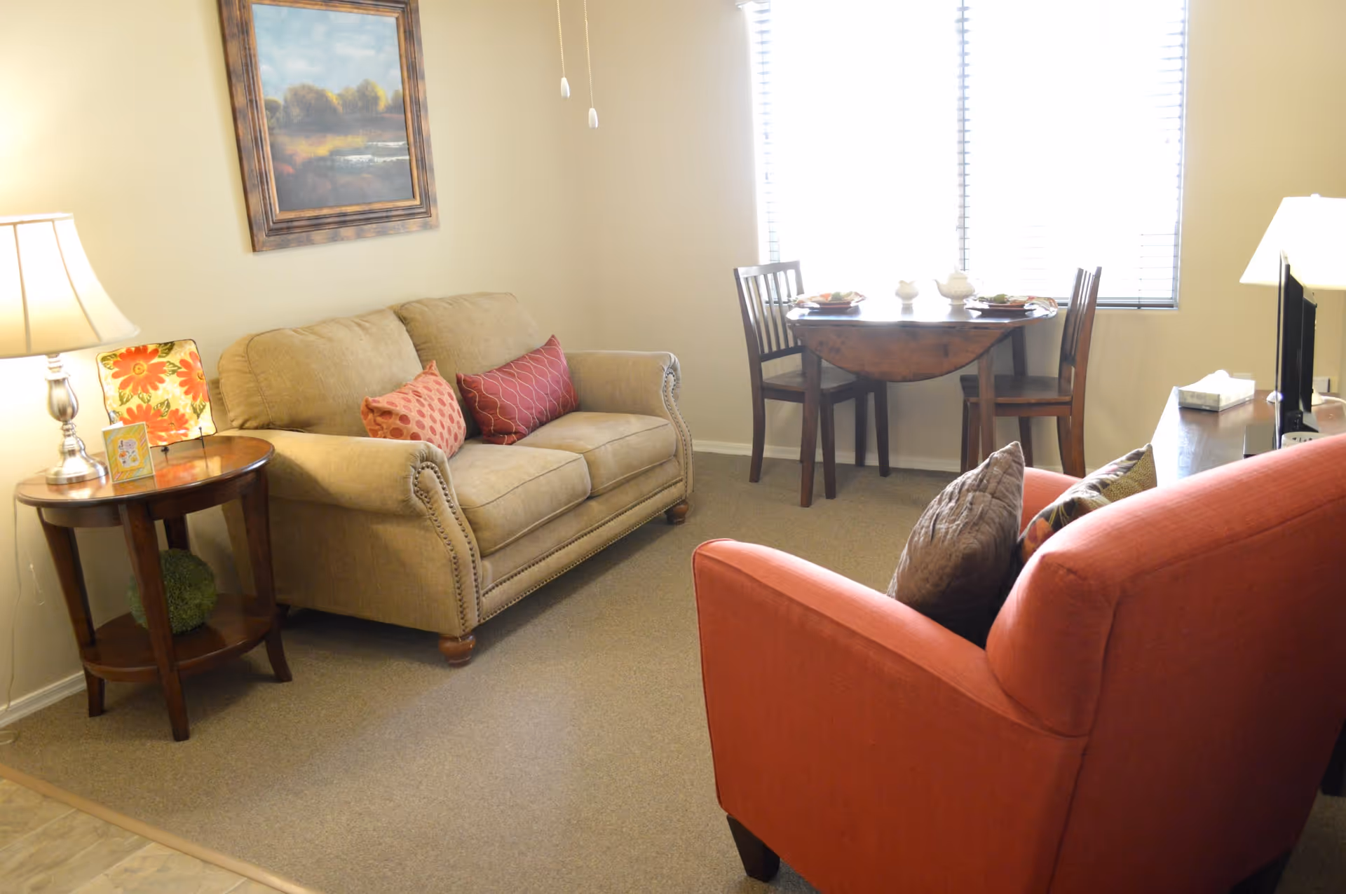 A cozy living room area with a beige loveseat adorned with two decorative pillows, a red armchair with pillows, a wooden side table with a lamp and decorative items, a small wooden dining table with two chairs near a window with blinds, and a framed landscape painting on the wall.
