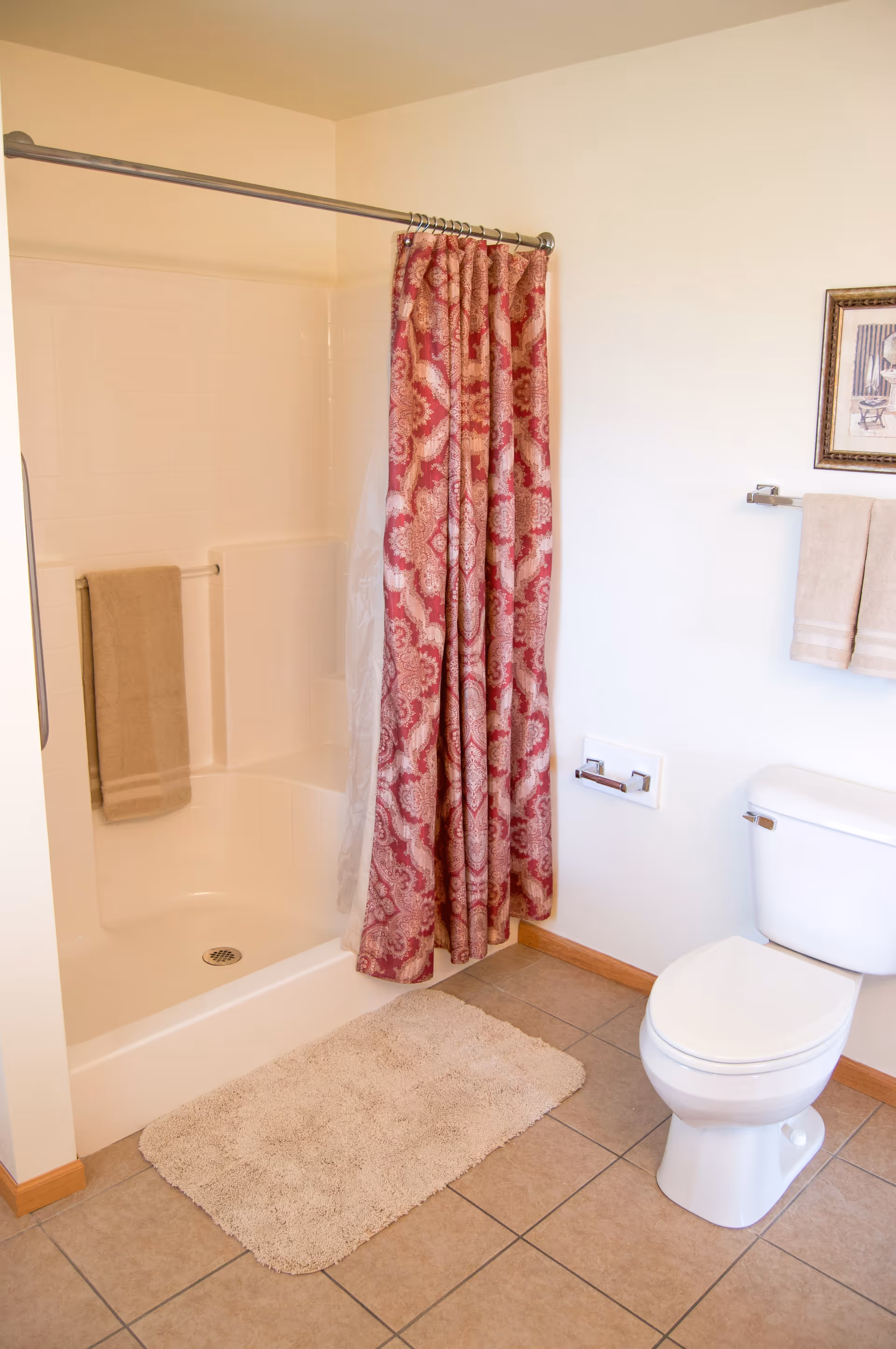 Bathroom with a shower-tub featuring a patterned red curtain, toilet, towel rack, and beige bath mat on tiled floor.
