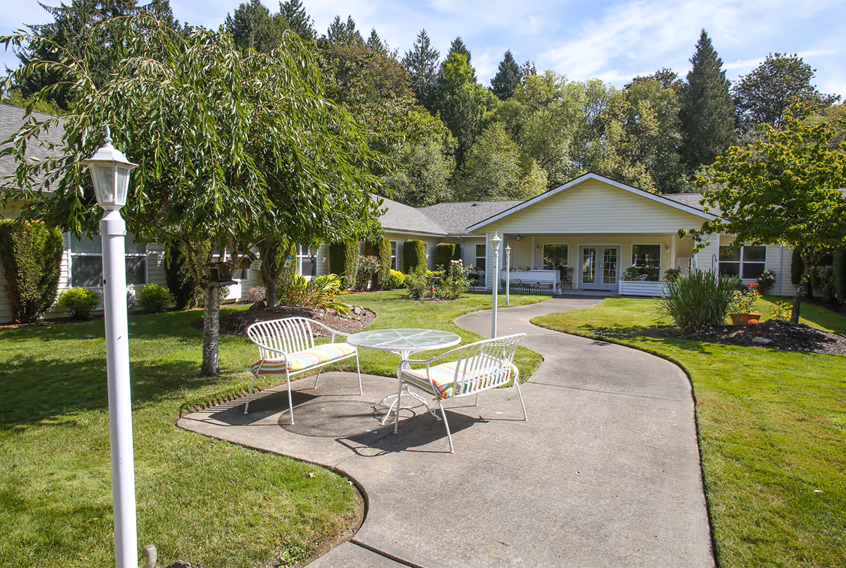 Outdoor seating area with two white metal benches with cushions and a round glass table on a concrete patio surrounded by green grass, trees, and shrubs. A paved walkway leads to a single-story building with a covered porch and large windows, set against a backdrop of tall trees under a partly cloudy sky.