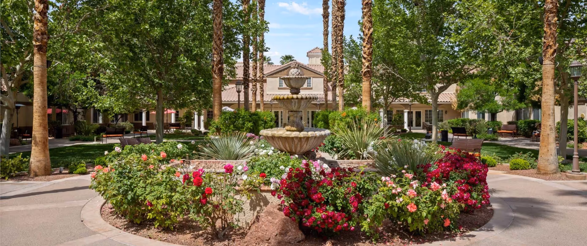 Sunny courtyard with a tiered fountain surrounded by flower beds, palm trees, benches and the facility building in the background.
