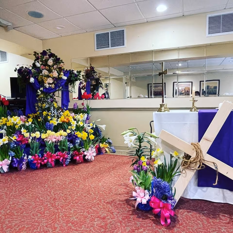 Indoor room decorated for a religious event with a large floral cross covered in purple cloth and surrounded by colorful flowers with pink ribbons. A wooden cross is leaning against a table draped in white and purple cloth, with two brass candle holders and a brass cross on top. The room has a red carpet and mirrored wall in the background reflecting framed pictures.