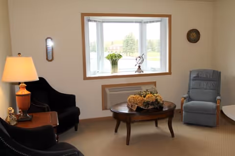 Cozy sitting area with upholstered armchairs, a coffee table, lamp, and a bay window overlooking greenery.
