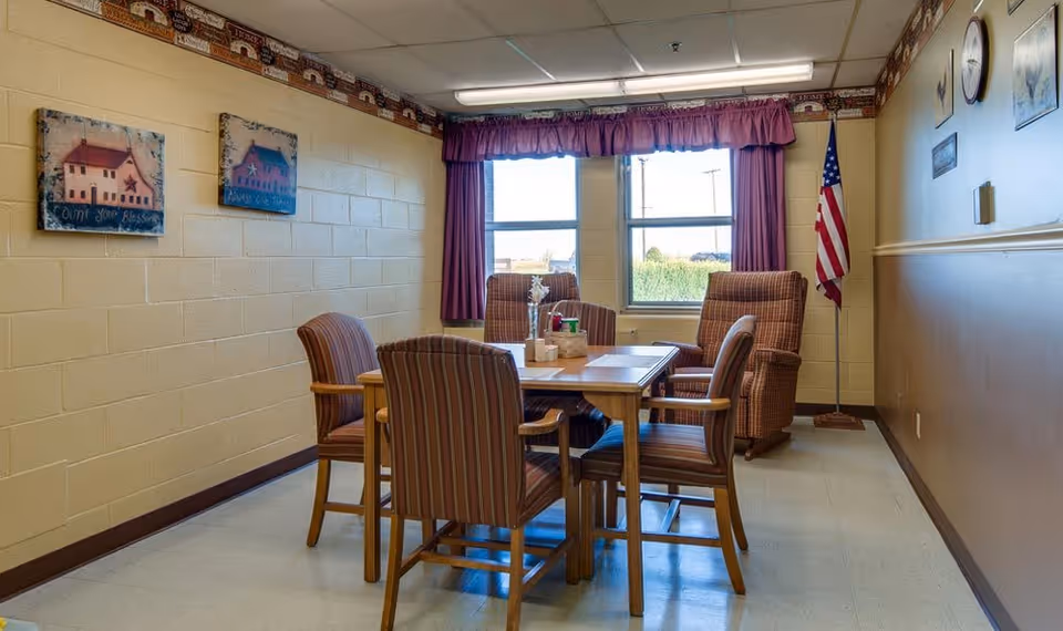A small dining area with a wooden table surrounded by four striped upholstered chairs and two cushioned armchairs near a window with purple curtains. The walls are painted beige with decorative paintings and an American flag in the corner.
