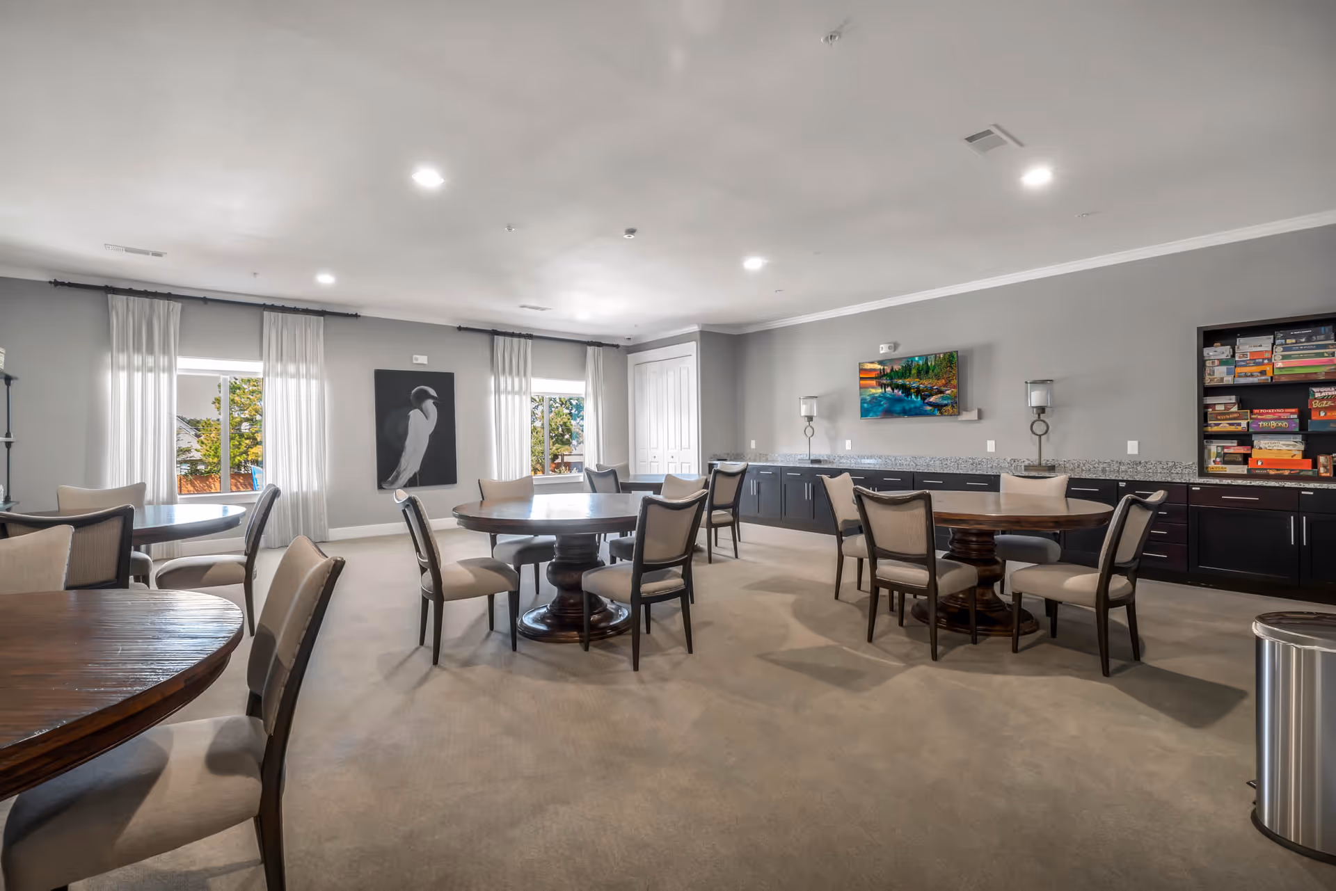 A spacious common room with round wooden tables and cushioned chairs arranged for group seating. The room has gray walls, two windows with white curtains, a large painting of a bird on one wall, and a flat-screen TV mounted on another wall above a long counter with cabinets. A shelf filled with board games is visible on the right side.