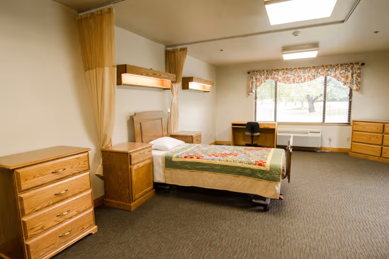 A spacious bedroom in a care facility with a single bed covered with a colorful quilt, wooden furniture including a dresser, nightstand, desk with a chair, and a chest of drawers. The room has a large window with floral curtains letting in natural light and beige walls with carpeted flooring.