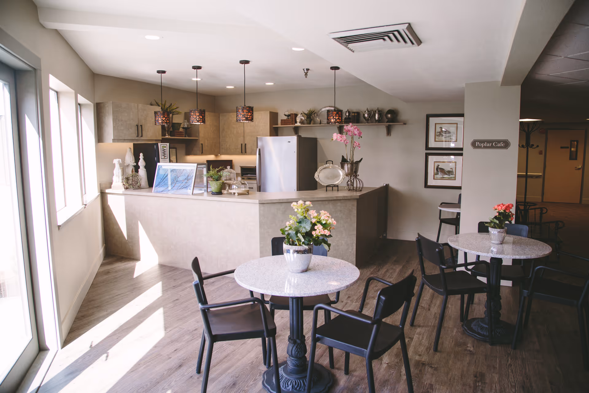 A bright and cozy cafe area inside Weatherly Inn Independent Senior Living featuring round tables with black chairs and potted flowers on each table. The background shows a kitchen counter with hanging pendant lights, cabinets, a refrigerator, and decorative items on shelves. A sign on the wall reads 'Poplar Cafe'.