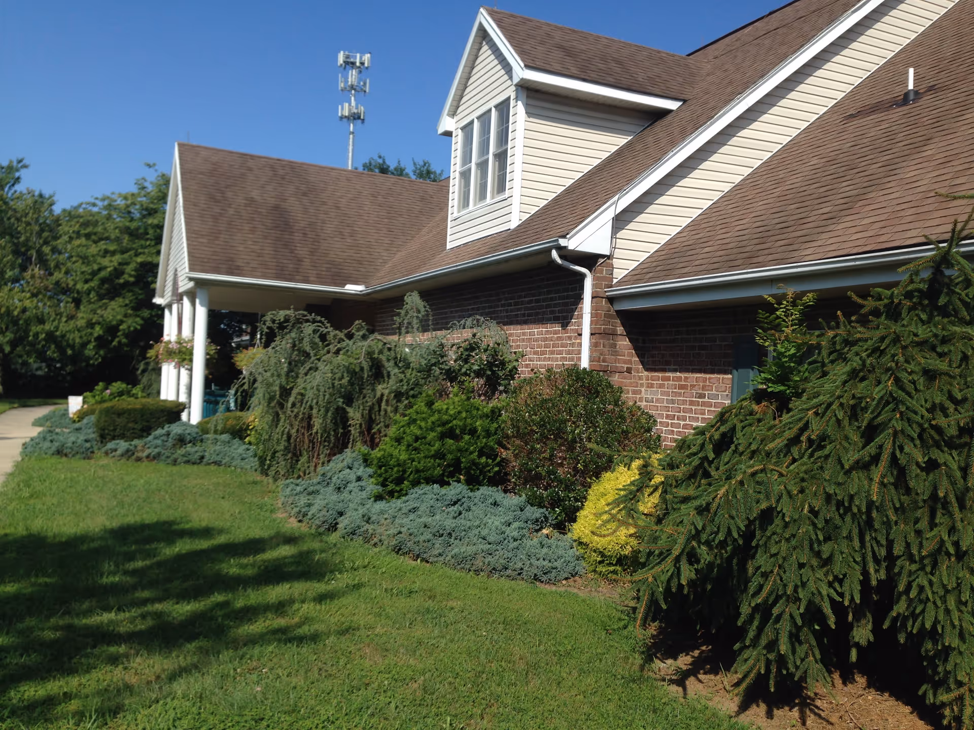 Exterior view of a senior living facility building with brick and beige siding, surrounded by well-maintained shrubs and greenery under a clear blue sky.