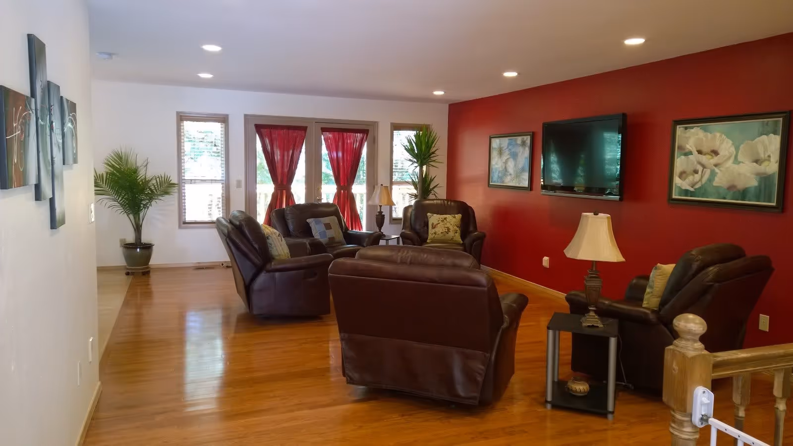 A cozy living room with wooden floors, four dark brown leather armchairs arranged around a central ottoman, a small side table with a lamp, two potted plants near windows with red curtains, and a red accent wall featuring a mounted flat-screen TV and two framed floral paintings.