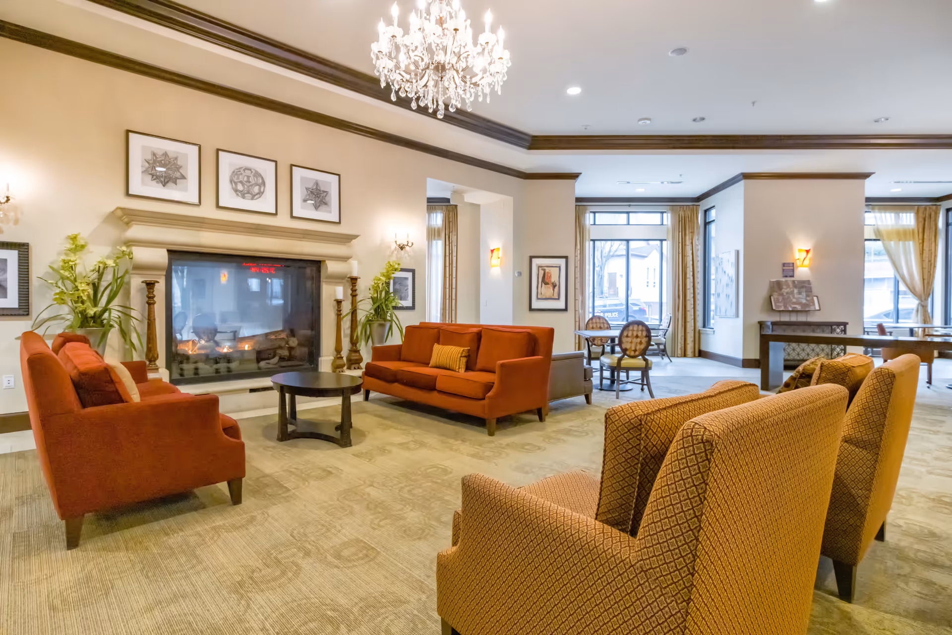 A spacious and elegant living room area in a senior living facility featuring a large fireplace with decorative framed art above it, two orange sofas, two patterned armchairs, a round coffee table, and large windows with curtains allowing natural light to fill the room. A chandelier hangs from the ceiling, and there are plants and additional seating areas visible in the background.