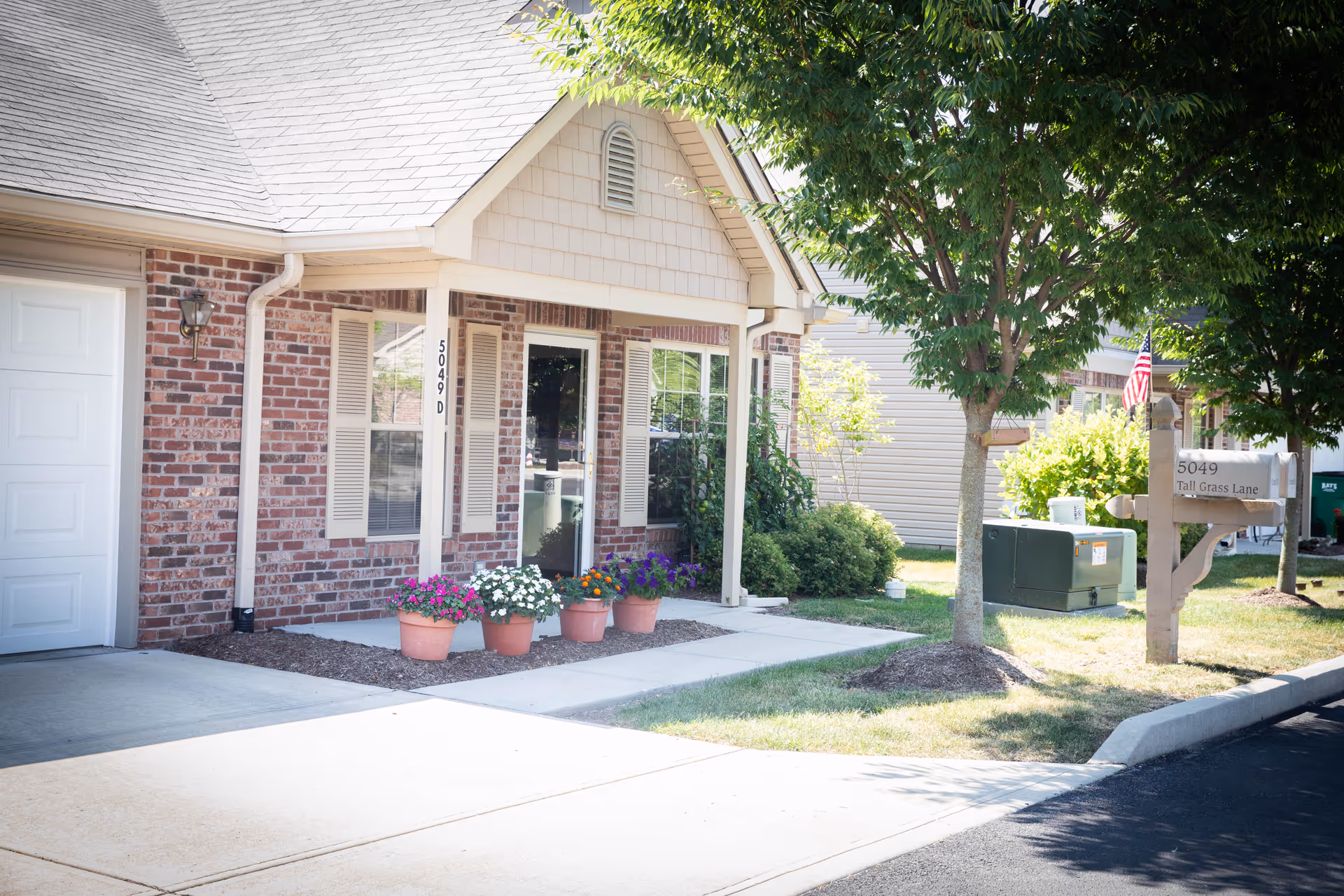 Exterior view of a brick and siding assisted living facility unit with a white garage door, a glass front door, and windows with shutters. There are four flower pots with colorful flowers arranged along the sidewalk leading to the entrance. A tree and mailbox with the address 5049 Tall Grass Lane are visible on the right side, along with some greenery and an American flag in the background.