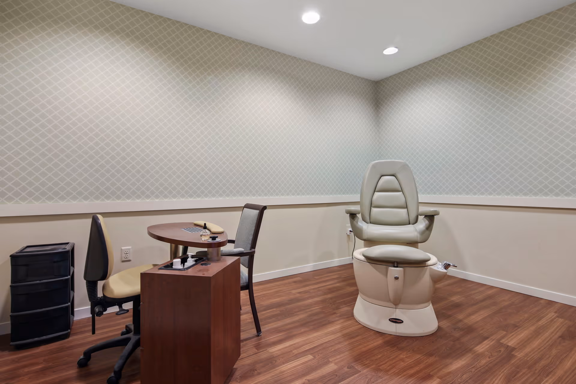 A small room with wood flooring and patterned wallpaper, featuring a beige pedicure spa chair, a small round table with a manicure station, and two chairs, one on each side of the table.