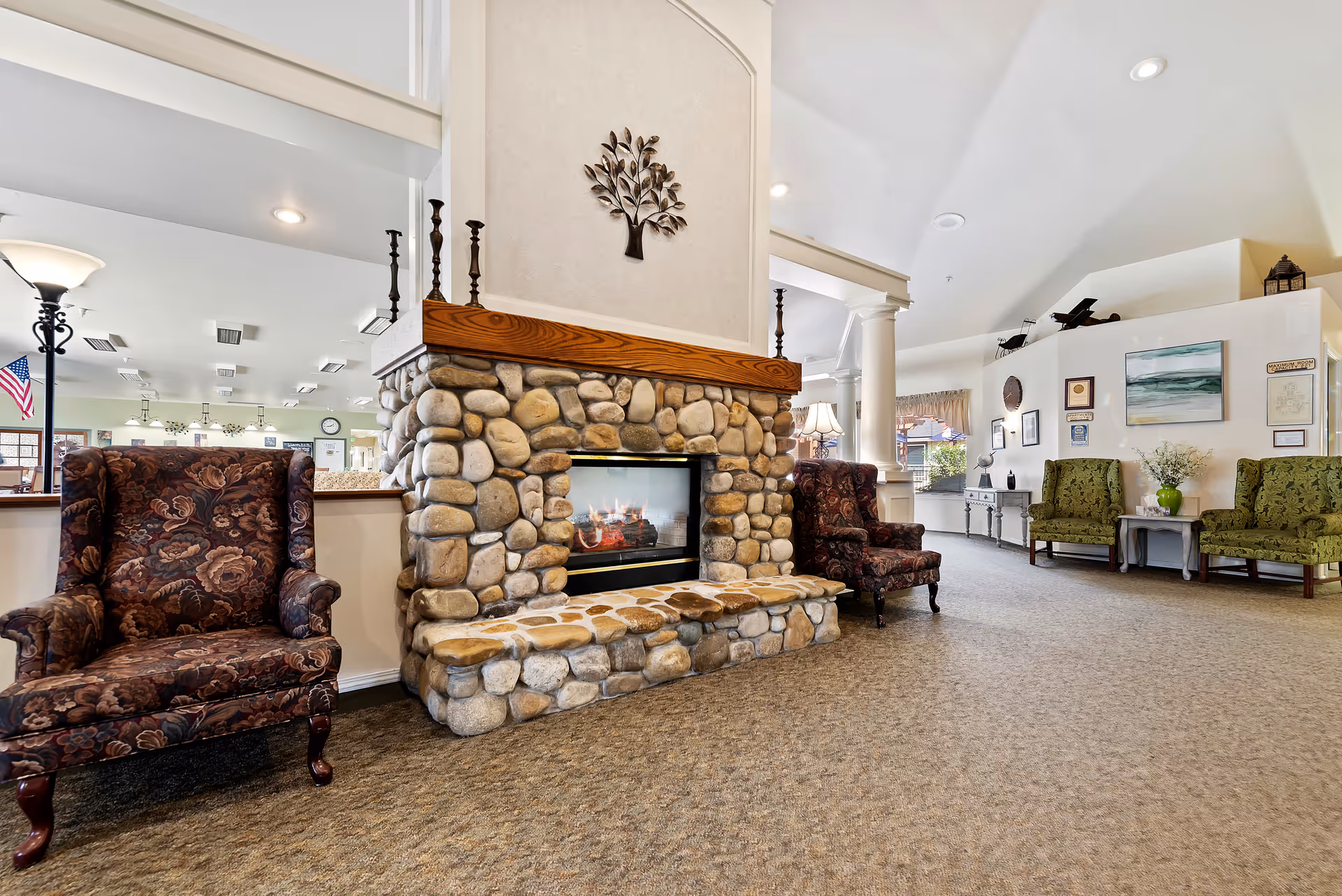 A cozy assisted living facility common area with a stone fireplace in the center, flanked by two patterned armchairs. The room has carpeted floors, high ceilings with recessed lighting, and additional seating with green upholstered chairs along the wall. Decorative items and framed pictures adorn the walls, creating a warm and inviting atmosphere.