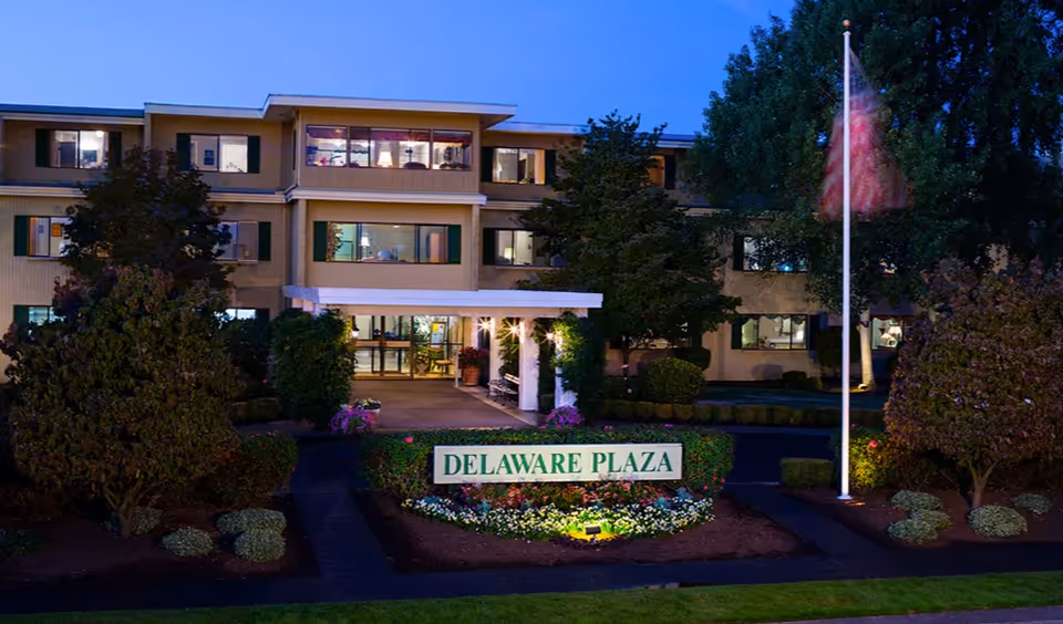 Exterior front view of Delaware Plaza Assisted Living Community building at dusk, with illuminated windows, landscaped garden, American flag on a flagpole, and a sign reading 'DELAWARE PLAZA' surrounded by flowers.