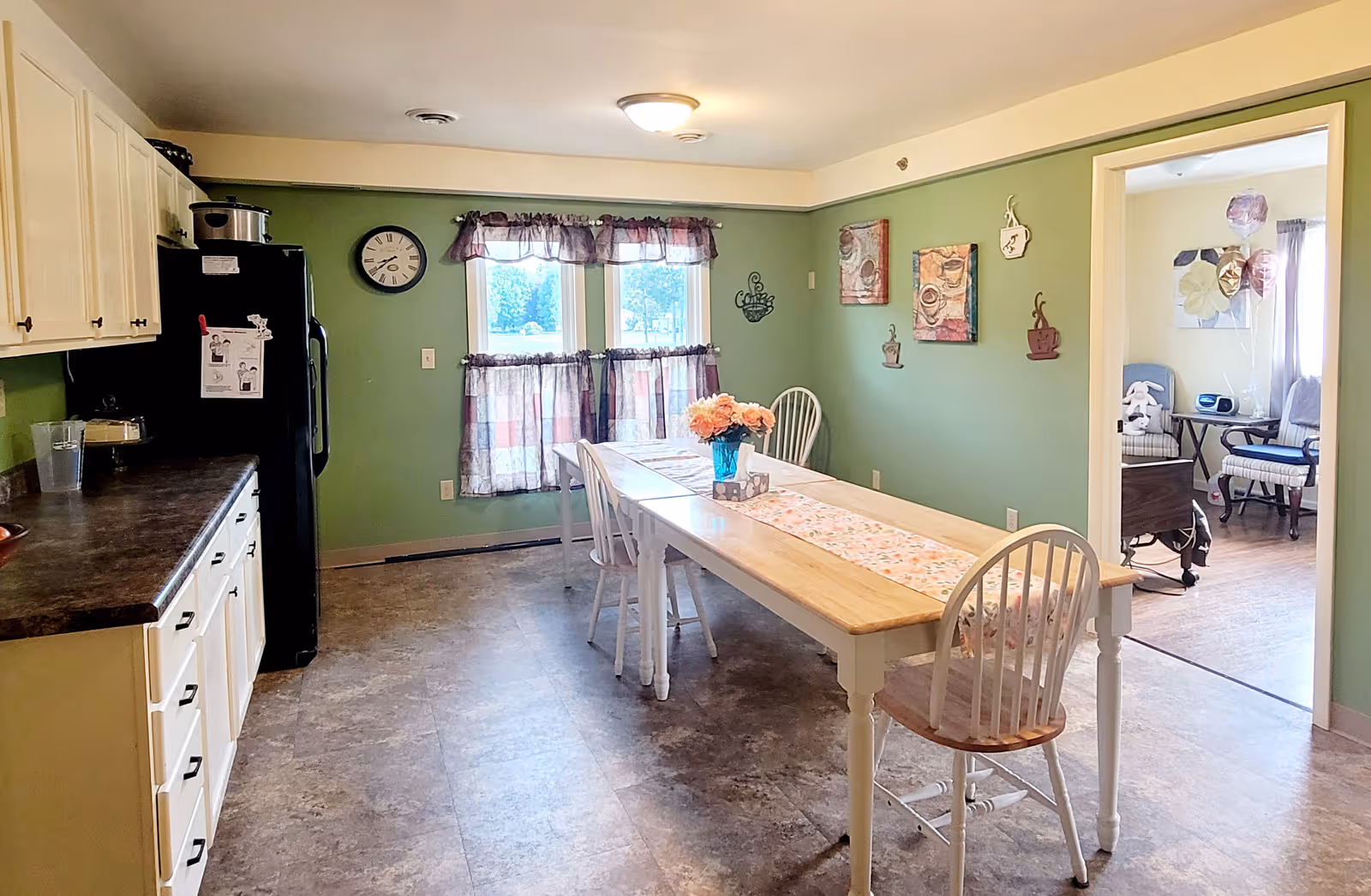 A dining room with green walls and a long wooden table with a floral runner and a vase of flowers in the center. There are four white chairs around the table. To the left, there is a kitchen area with white cabinets, a dark countertop, and a black refrigerator. A clock is mounted on the wall above the refrigerator. Two windows with purple curtains let in natural light. Through an open doorway, a room with chairs and a small table is visible.