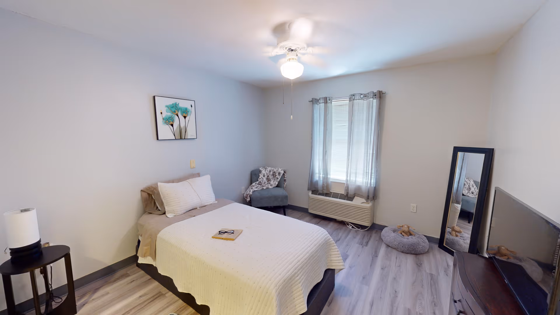 A simple, clean bedroom with a single bed covered in a white quilt, a small round side table with a lamp, a gray armchair with a patterned throw, a window with sheer curtains, a full-length mirror leaning against the wall, a TV on a dark wooden dresser, and a pet bed on the floor.