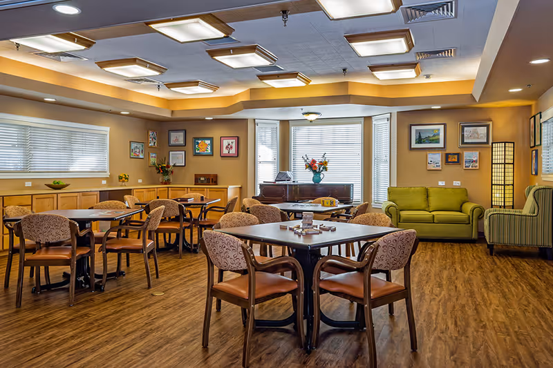Bright multipurpose common room with tables and chairs, a green sofa, piano, and framed artwork on the walls.
