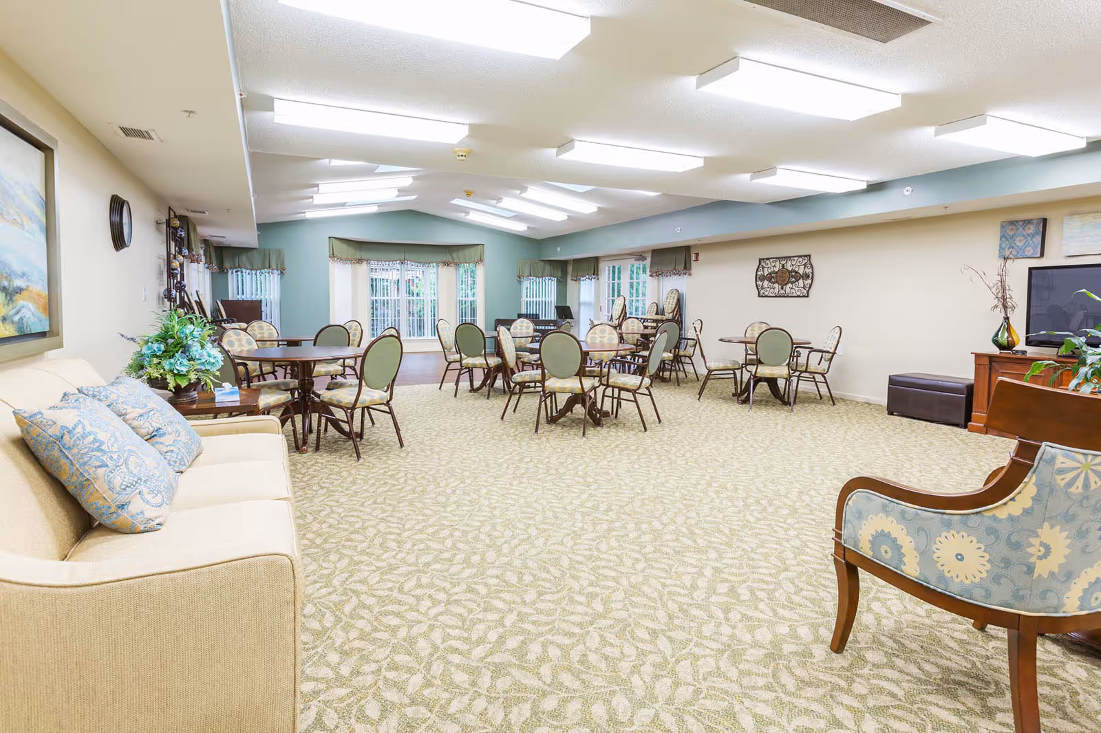 A spacious common area with multiple round tables and chairs arranged throughout the room. The room has a patterned carpet, a beige sofa with blue patterned pillows on the left, and a wooden armchair with blue floral upholstery on the right. The walls are light-colored with some decorative artwork and a large flat-screen TV on a wooden stand. Large windows with green valances allow natural light to enter the room.