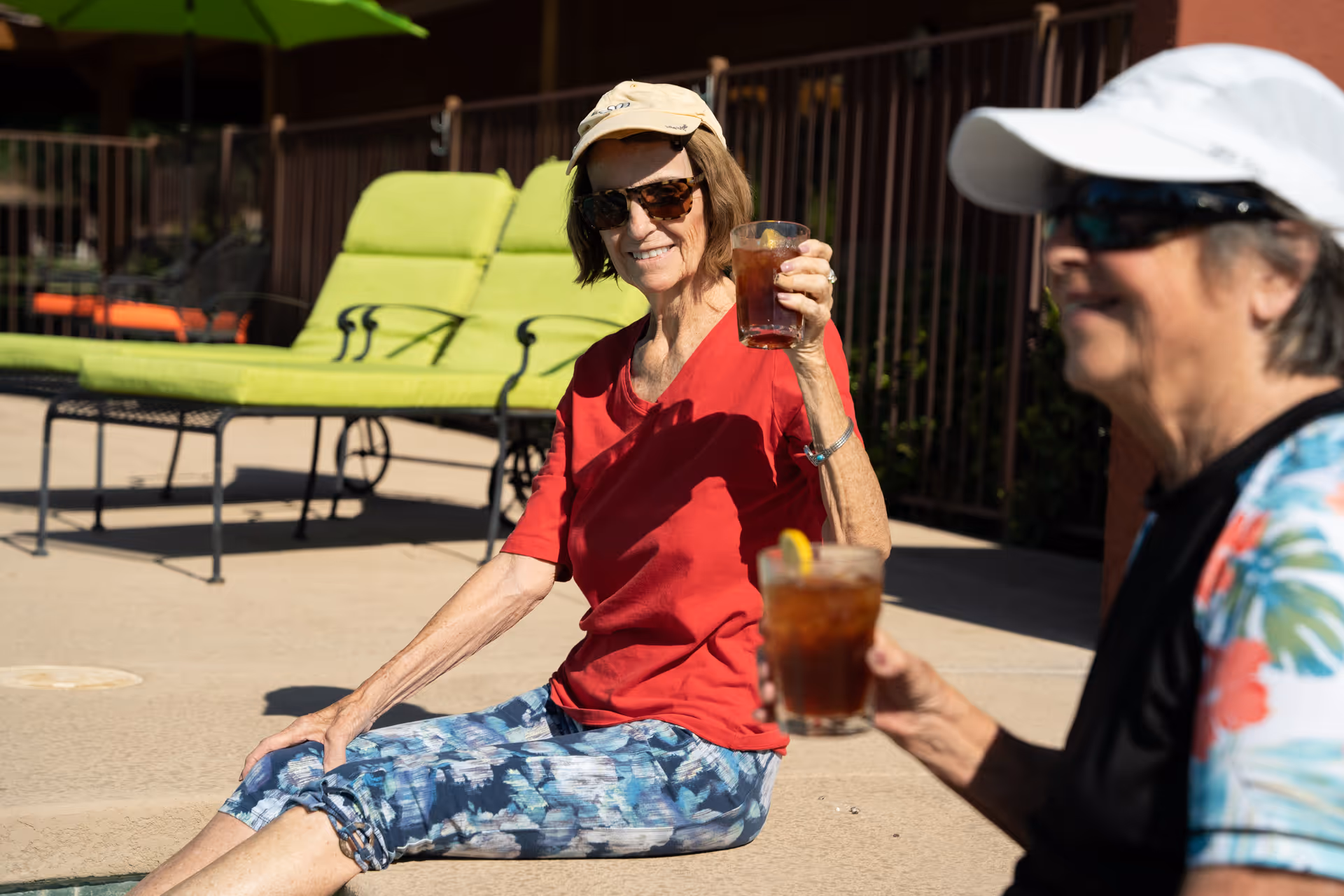 Two elderly women sitting by a poolside, each holding a glass of iced tea. One woman is wearing a red shirt, patterned pants, sunglasses, and a beige cap, while the other woman is wearing a floral shirt, sunglasses, and a white cap. Green lounge chairs and a fence are visible in the background.