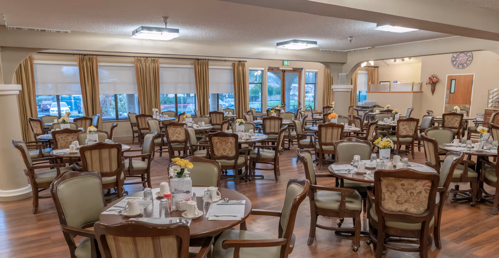A spacious dining room with multiple round tables set with white cups, glasses, napkins, and silverware. Each table has a small flower arrangement and a menu card. The room features large windows with beige curtains, wooden flooring, and a neutral color scheme. There are several chairs around each table, and the room is well-lit with ceiling lights.
