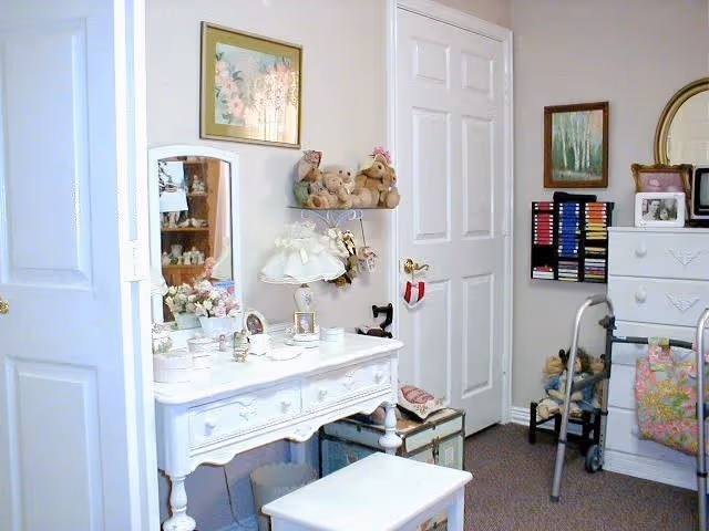 A cozy room with white furniture including a vanity table with a mirror, a small bench, and a chest of drawers. The vanity table has various decorative items such as a lamp, framed photos, and small containers. There are teddy bears on a wall shelf above the vanity. A walker is positioned near the chest of drawers, and a rack with colorful items is mounted on the wall. The room has light-colored walls with framed artwork and a carpeted floor.