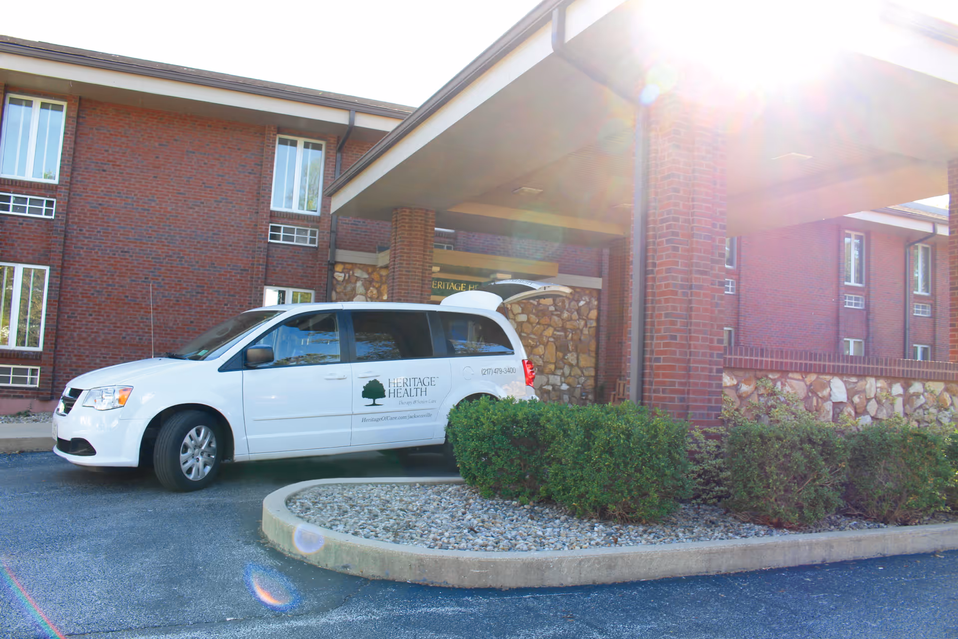A white van with Heritage Health logo parked under a covered entrance of a brick building with stone accents and several windows. There are bushes and a small landscaped area with rocks in front of the building.