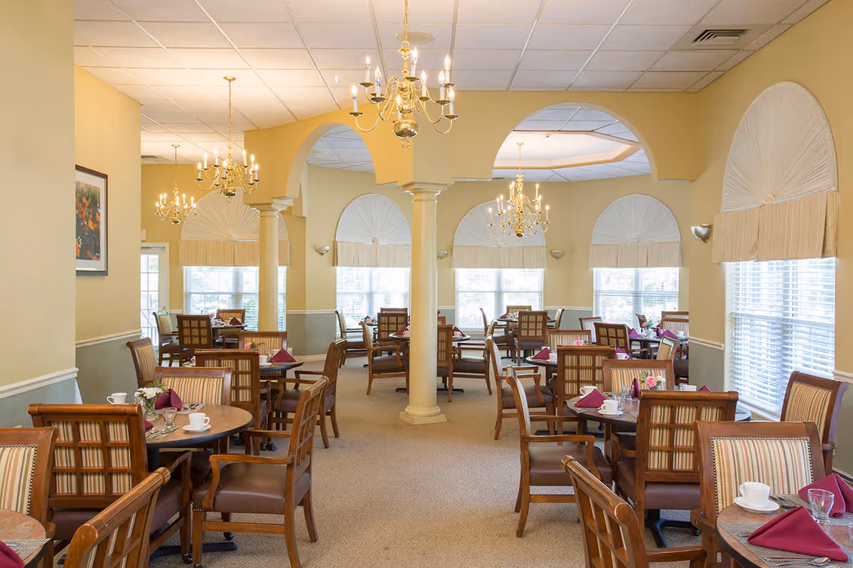 A bright and elegant dining room with multiple round wooden tables and cushioned chairs. Each table is set with white cups, glasses, silverware, and folded burgundy napkins. The room features large windows with decorative white valances, beige walls, carpeted floors, and several gold chandeliers hanging from the ceiling. There are architectural columns and arches dividing the space.