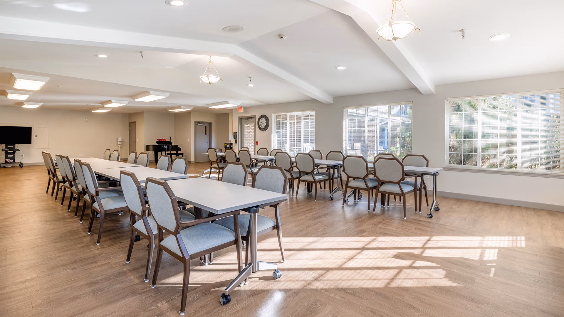 A bright and spacious dining room with multiple tables and chairs arranged neatly. Large windows allow natural light to fill the room, and there is a piano and a wall-mounted TV in the background. The room has wooden flooring and white walls with ceiling lights.