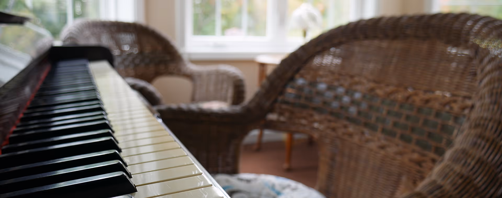 Close-up of a piano keyboard with wicker chairs and a sunlit window in the background.