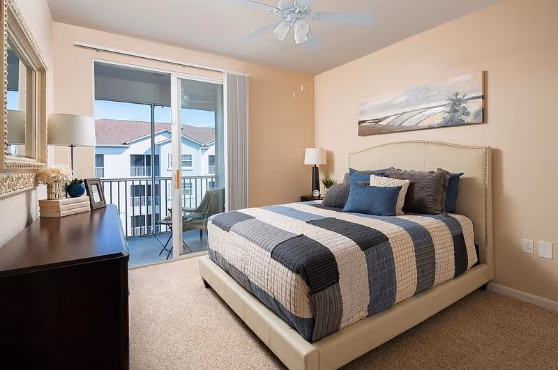 A neatly made bed with a striped blue, gray, and beige quilt in a bedroom. The room features a ceiling fan, a dresser with a lamp, framed pictures, and decorative items. Sliding glass doors lead to a balcony with outdoor seating and a view of neighboring buildings.