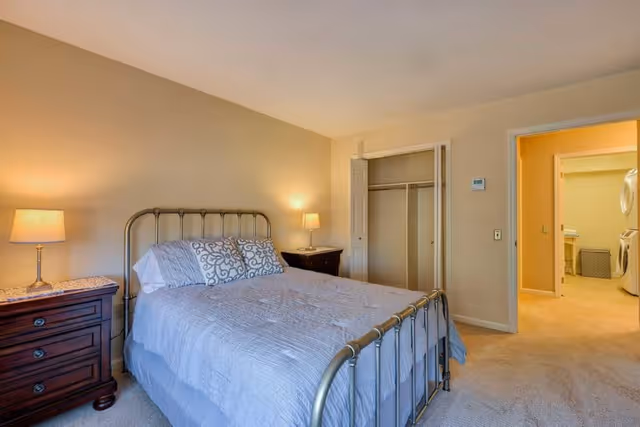 A bedroom with a metal frame bed covered with a gray quilt and decorative pillows. There are two wooden nightstands with lamps on either side of the bed. An open closet with sliding doors is visible on the right wall, and an open doorway leads to a laundry area with a washer and dryer. The room has beige walls and carpeted flooring.