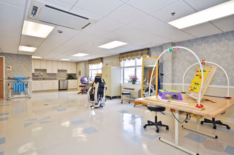 A spacious therapy or rehabilitation room with exercise equipment, therapy tools on a table, and a kitchenette area in the background. The room has large windows with valances, patterned wallpaper, and a shiny tiled floor.