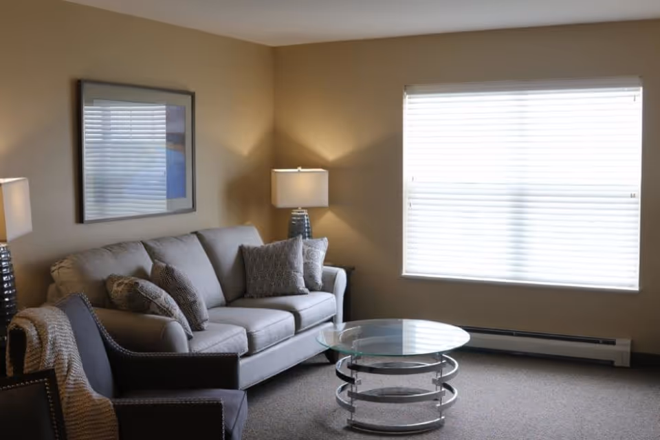 Well-lit living room with a gray sofa, armchair, glass coffee table, lamps, and a large window with blinds.