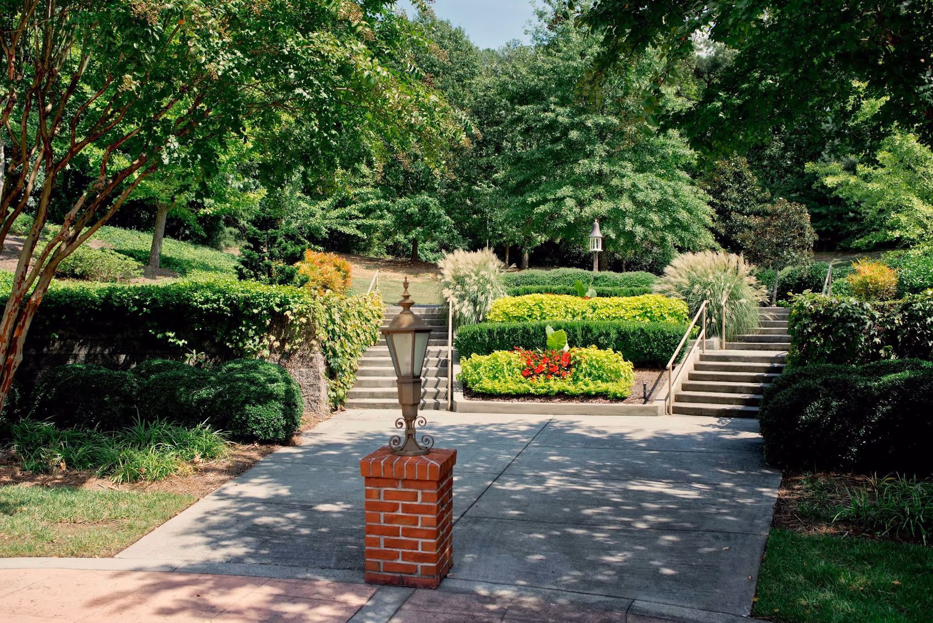 A landscaped outdoor area with a concrete pathway leading to two staircases on either side. The area is surrounded by green bushes, trees, and colorful flower beds. A decorative lamp post on a brick base is centered in the foreground.
