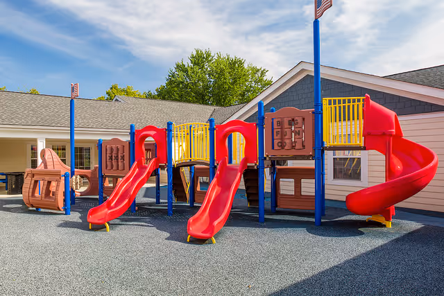 Outdoor playground area with multiple red slides, climbing structures, and play panels set on a rubberized safety surface, adjacent to a building with beige siding and a gray roof under a partly cloudy sky.