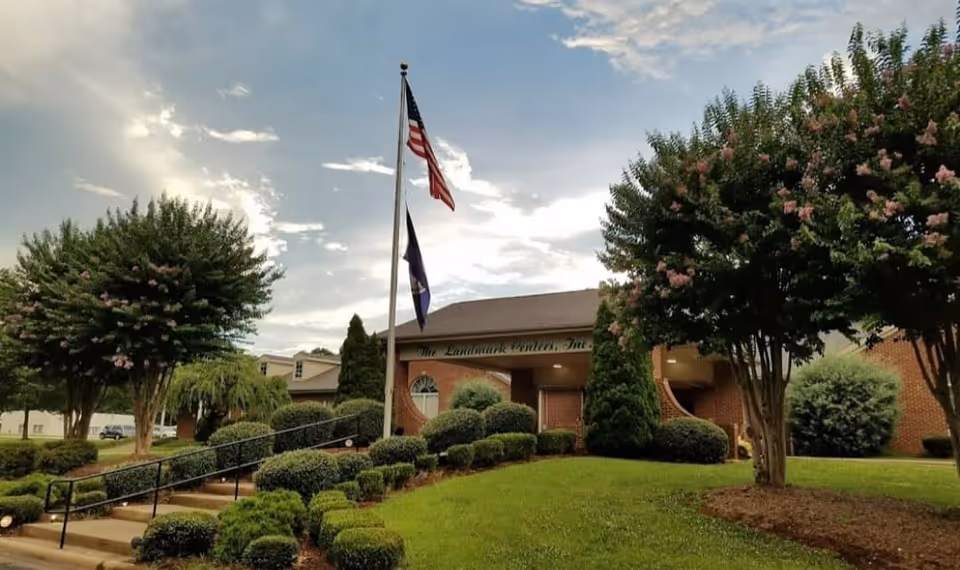 Exterior view of The Landmark Center building with a well-maintained lawn, bushes, and trees. Two flagpoles display the American flag and another flag in front of the building under a partly cloudy sky.