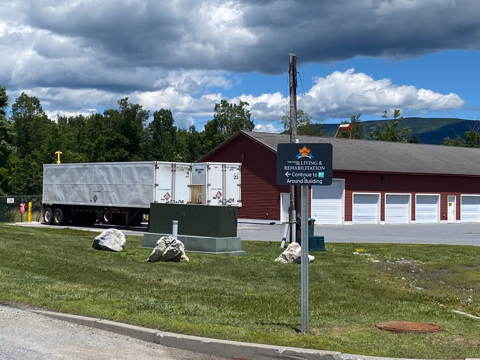 Red rehabilitation building with multiple garage doors, a parked white trailer, utility boxes, rocks and a sign for The Center for Living & Rehabilitation on a grassy lawn under a cloudy sky.