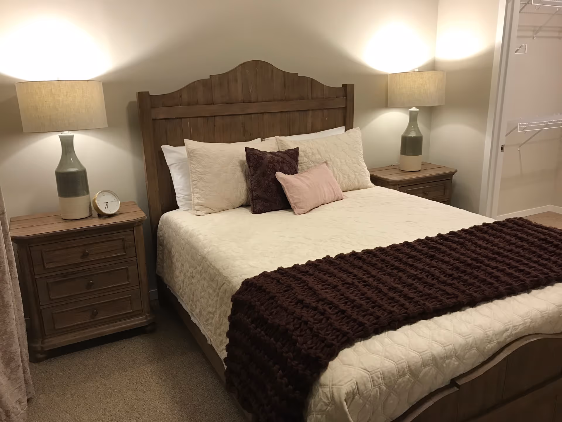 A cozy bedroom featuring a wooden bed frame with a cream-colored quilt and multiple pillows, including a dark purple and a light pink one. There are matching wooden nightstands on each side of the bed, each with a lamp and one with a small clock. The room has beige walls and carpet, and an open closet with wire shelving is visible on the right side.