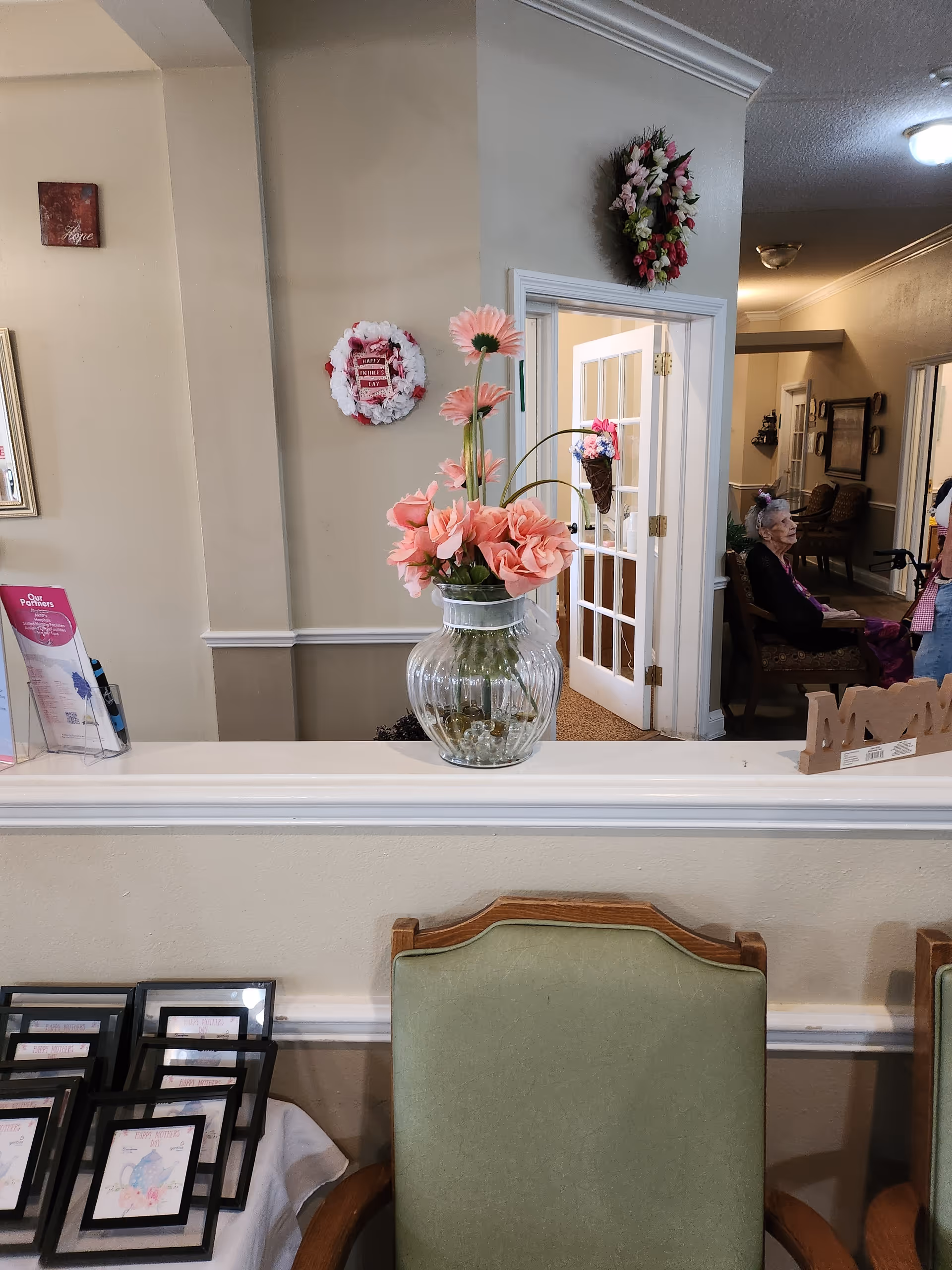 Interior view of a senior living facility reception or common area with a glass vase holding pink flowers on a white ledge. Behind the ledge, there is a hallway with an elderly woman sitting on a chair and a person standing nearby. The walls are decorated with floral wreaths and framed pictures. A green cushioned chair is visible in the foreground, along with a table holding several framed items.